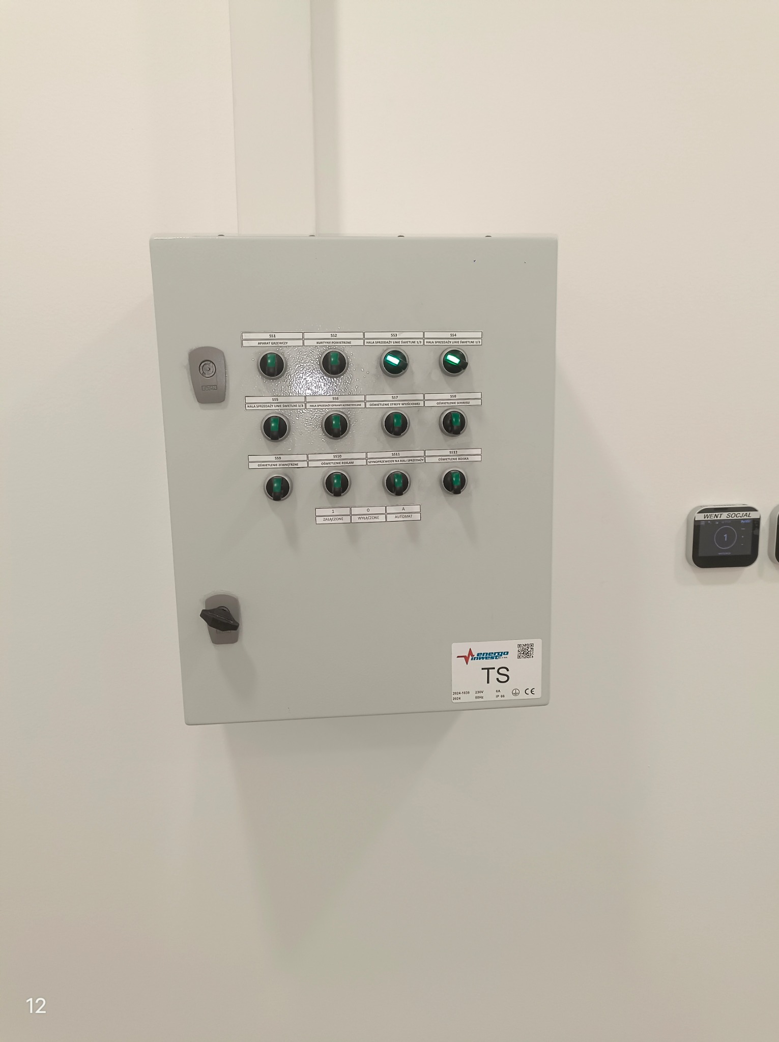 Szara szafa sterownicza z zielonymi diodami sygnalizacyjnymi, opisana etykietami z nazwami urządzeń i funkcji, zamontowana na białej ścianie, z widocznym logo firmy i parametrami technicznymi.