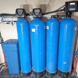 WODEKSPERT - Cztery niebieskie kolumny filtracyjne z panelami sterującymi, obok srebrny sterylizator UV i niebieski zbiornik, tworzące system uzdatniania wody w pomieszczeniu gospodarczym.