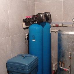 WODEKSPERT - Zainstalowany system do uzdatniania wody z dwoma niebieskimi filtrami, niebieskim zbiornikiem solanki i metalowym hydroforem w pomieszczeniu wyłożonym szarymi płytkami.
