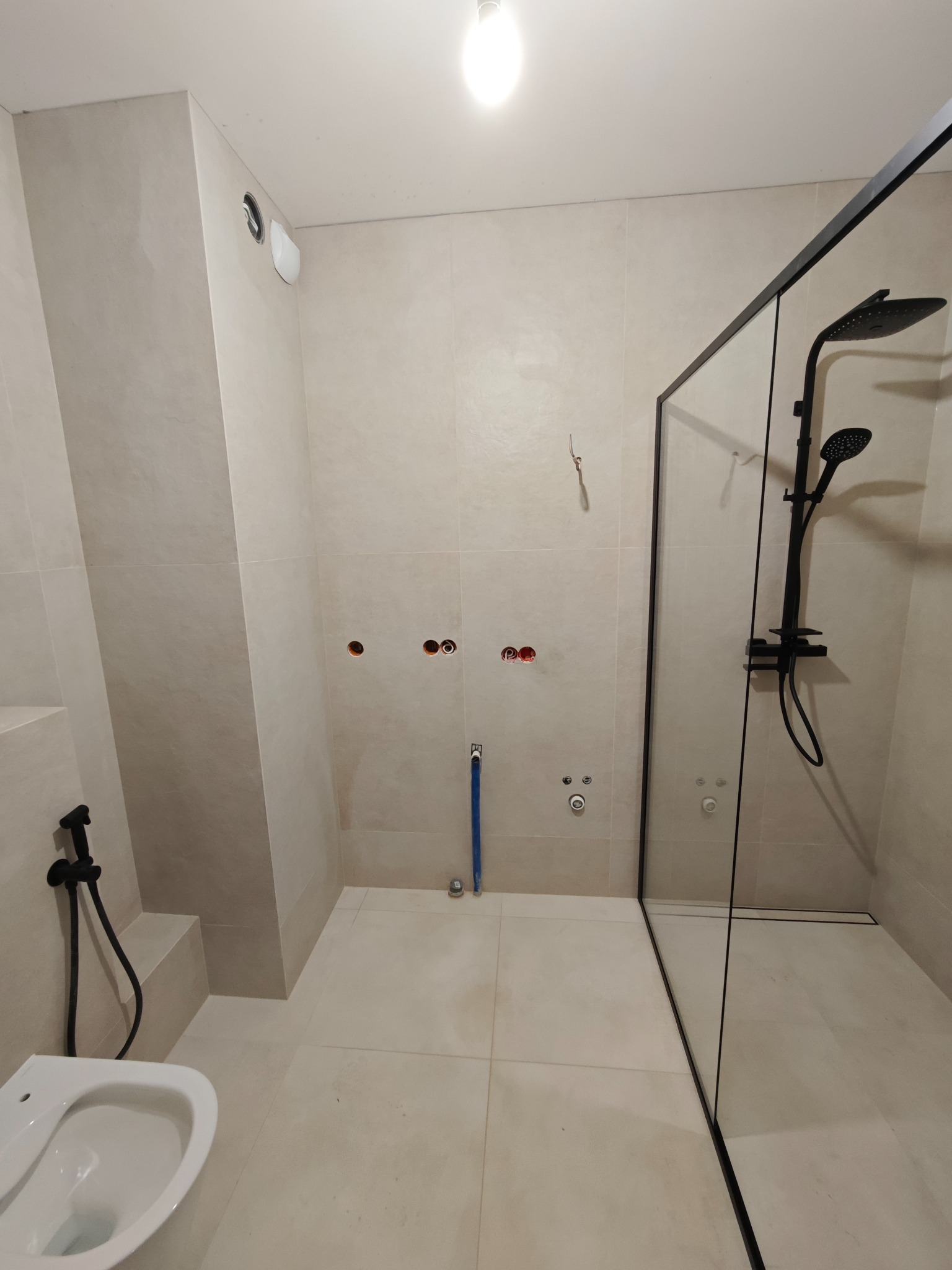Jasna łazienka w trakcie wykańczania: beżowe płytki, czarna armatura (deszczownica, bidetka), szklana kabina walk-in z czarną ramą. Widoczne przygotowane punkty instalacji.