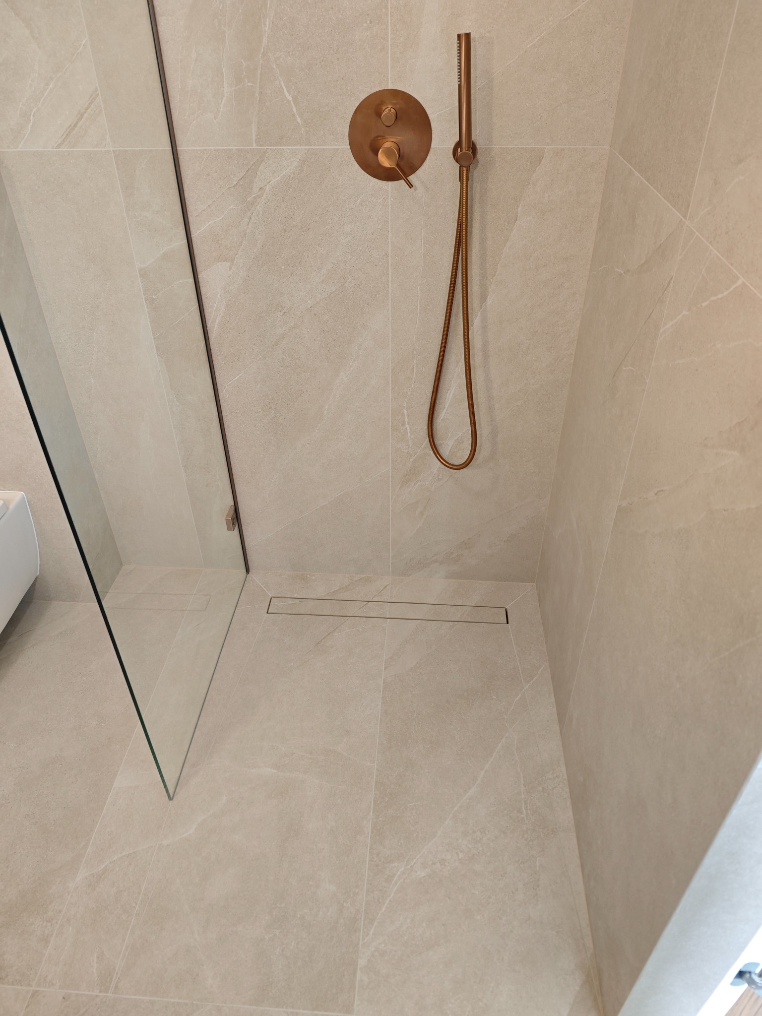 Beżowa kabina prysznicowa z odpływem liniowym i miedzianą armaturą, wykończona płytkami imitującymi kamień. Minimalistyczny design z elementami luksusu.