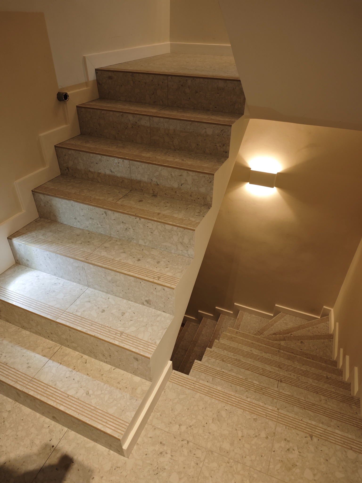 Wykończone schody z terazzo, z antypoślizgowymi paskami na krawędziach stopni. Widok z góry, oświetlenie lampą ścienną. Estetyczne wykończenie klatki schodowej.