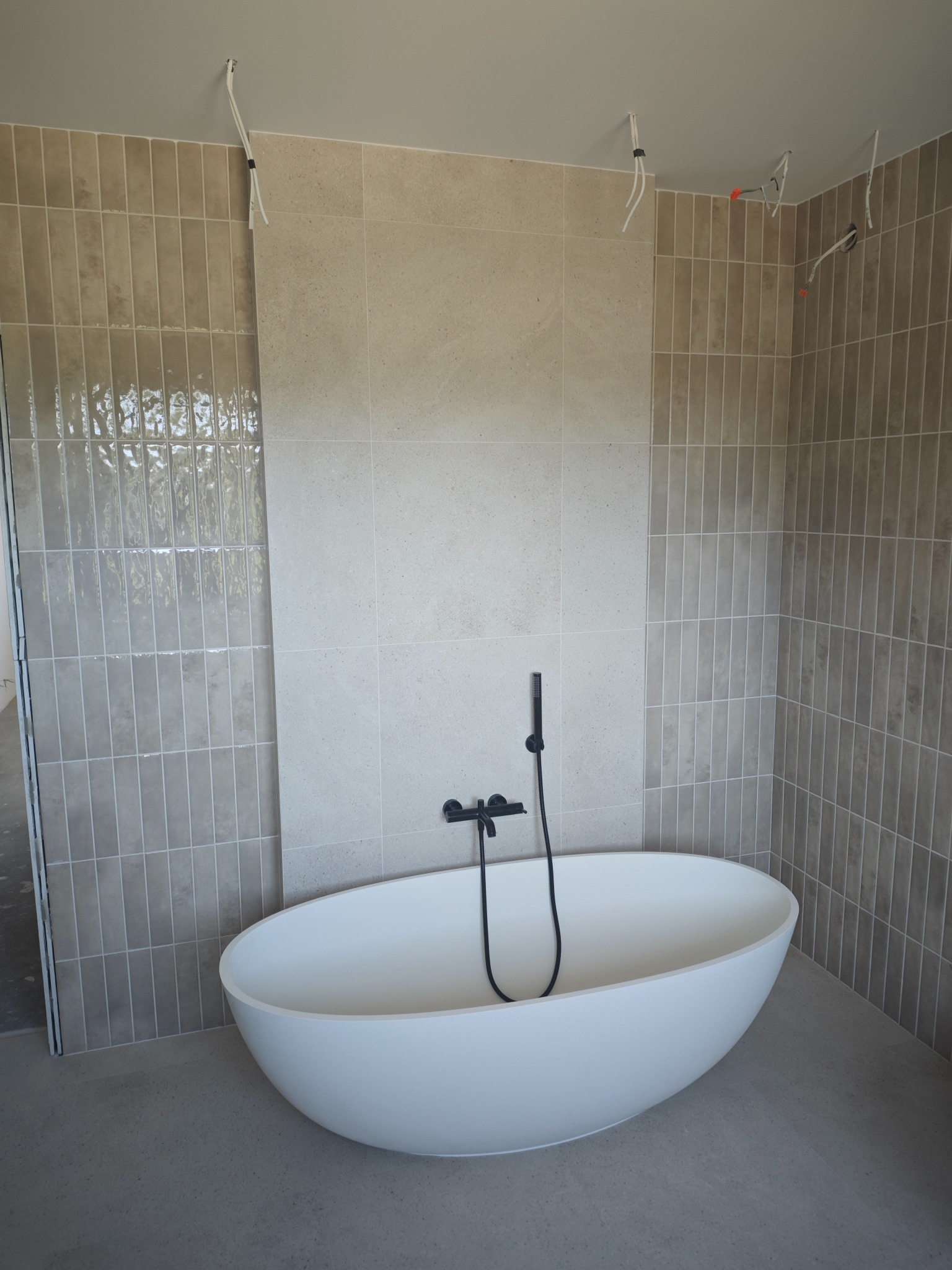 Nowoczesna łazienka w trakcie wykończenia: owalna wanna wolnostojąca, beżowe płytki na ścianach, widoczne przewody elektryczne. Minimalistyczny design.