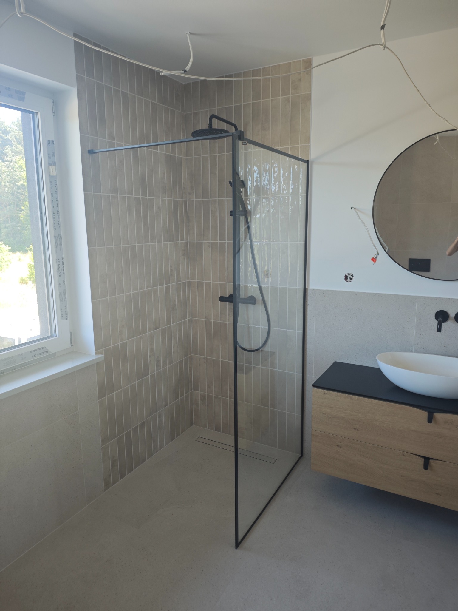 Nowoczesna łazienka z kabiną prysznicową typu walk-in z czarnymi profilami, umywalką nablatową i okrągłym lustrem. Beżowe płytki na ścianach i podłodze.