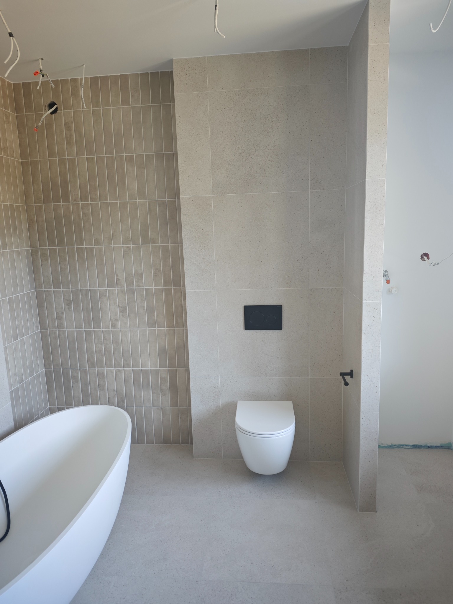 Nowoczesna łazienka w beżowych płytkach z wolnostojącą wanną i podwieszaną toaletą. Widoczne niedokończone instalacje elektryczne.