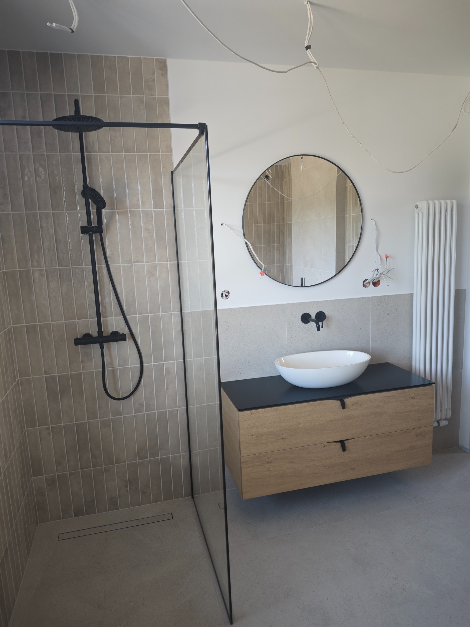 Nowoczesna łazienka z kabiną prysznicową typu walk-in, okrągłym lustrem i wiszącą szafką z umywalką. Beżowe płytki na ścianach i podłodze. Widoczne niedokończone podłączenia elektryczne.