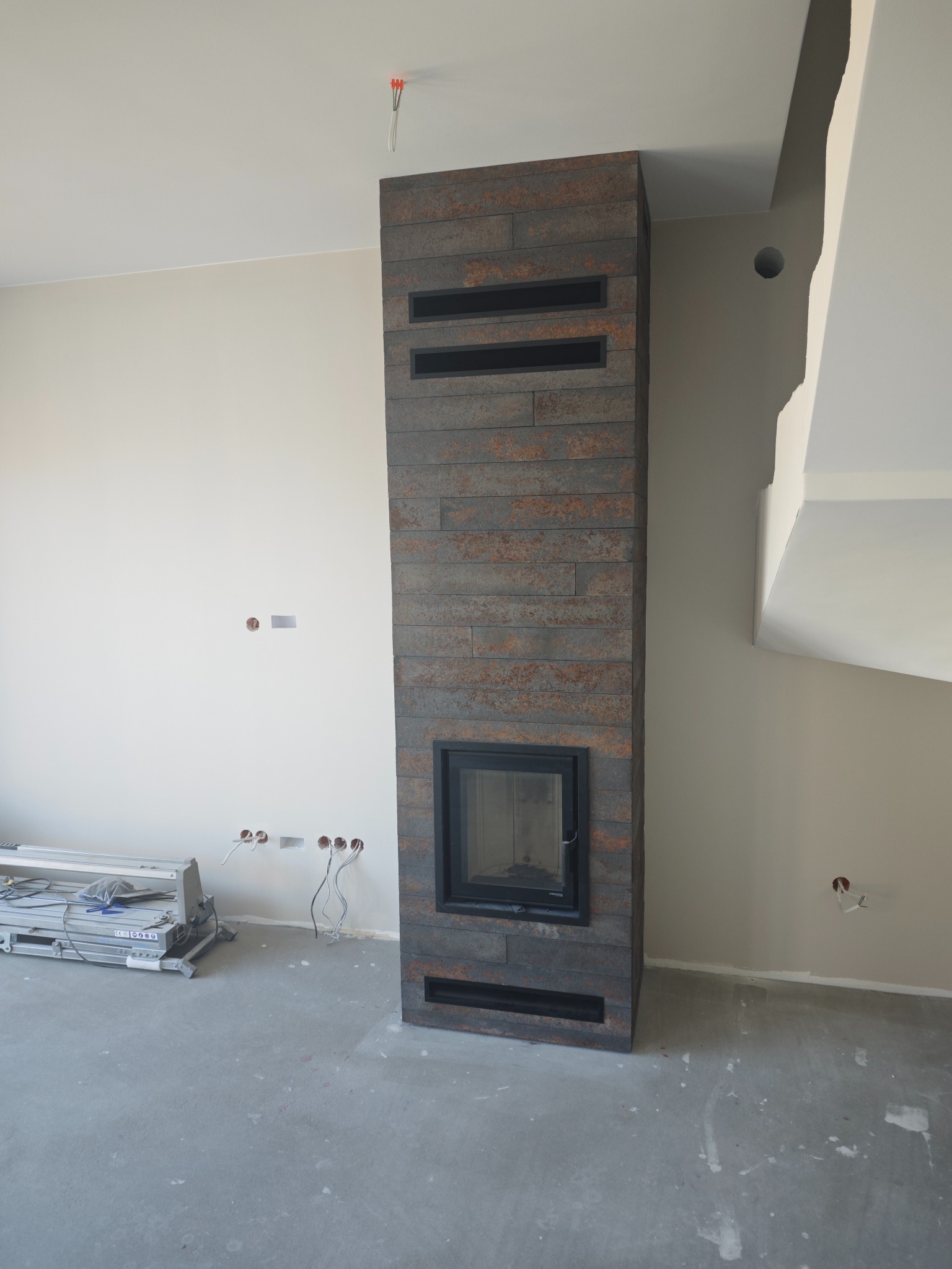 Obudowa kominka z płytek imitujących rdzę, z czarną wkładką. Widoczne niedokończone ściany i podłoga, sugerujące etap wykończenia wnętrza. Na podłodze rozłożona drabina.