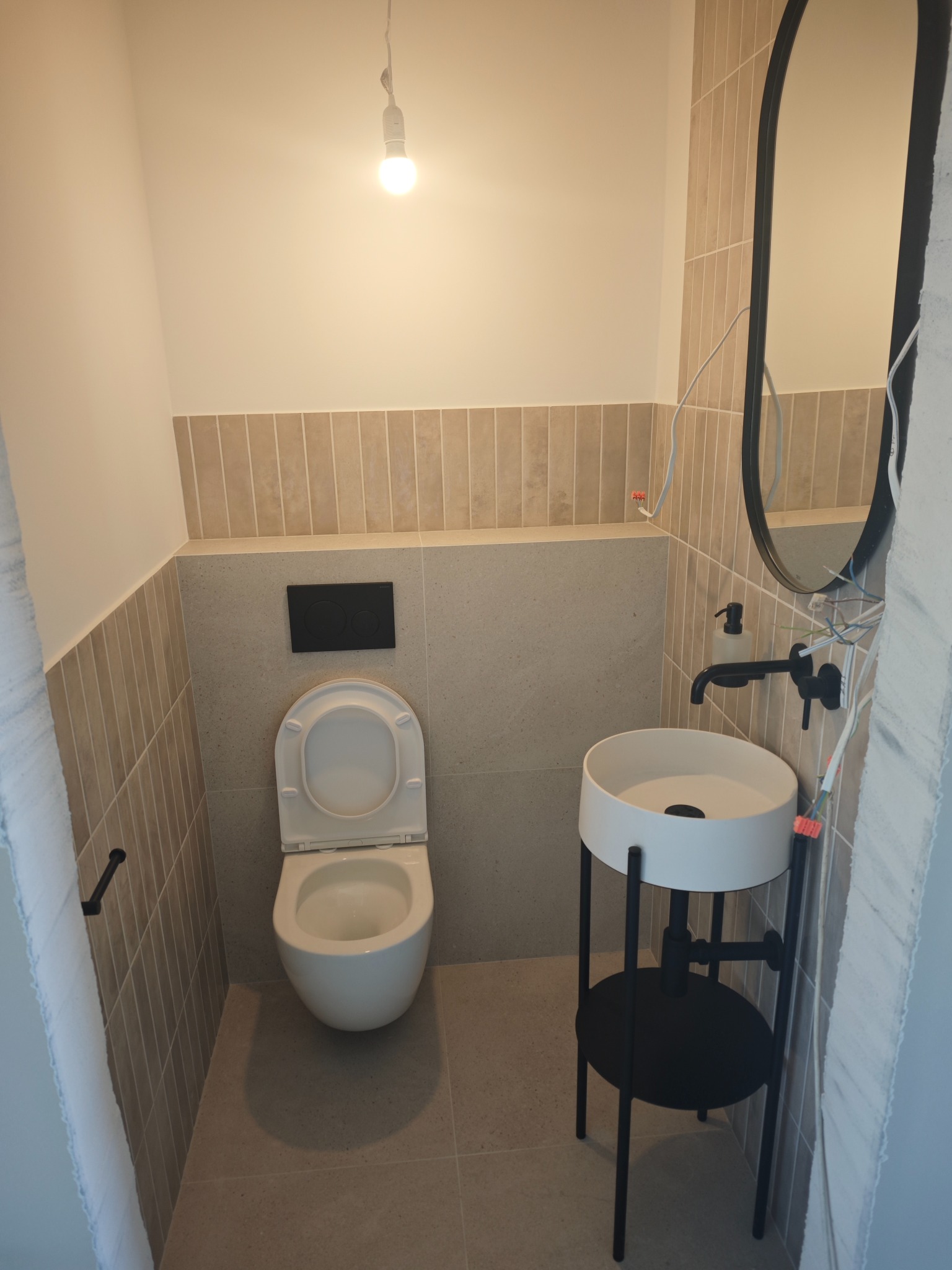 Nowoczesna toaleta z umywalką na metalowym stelażu i owalnym lustrem. Beżowe płytki na ścianach i podłodze. Minimalistyczny design, surowy styl wykończenia.