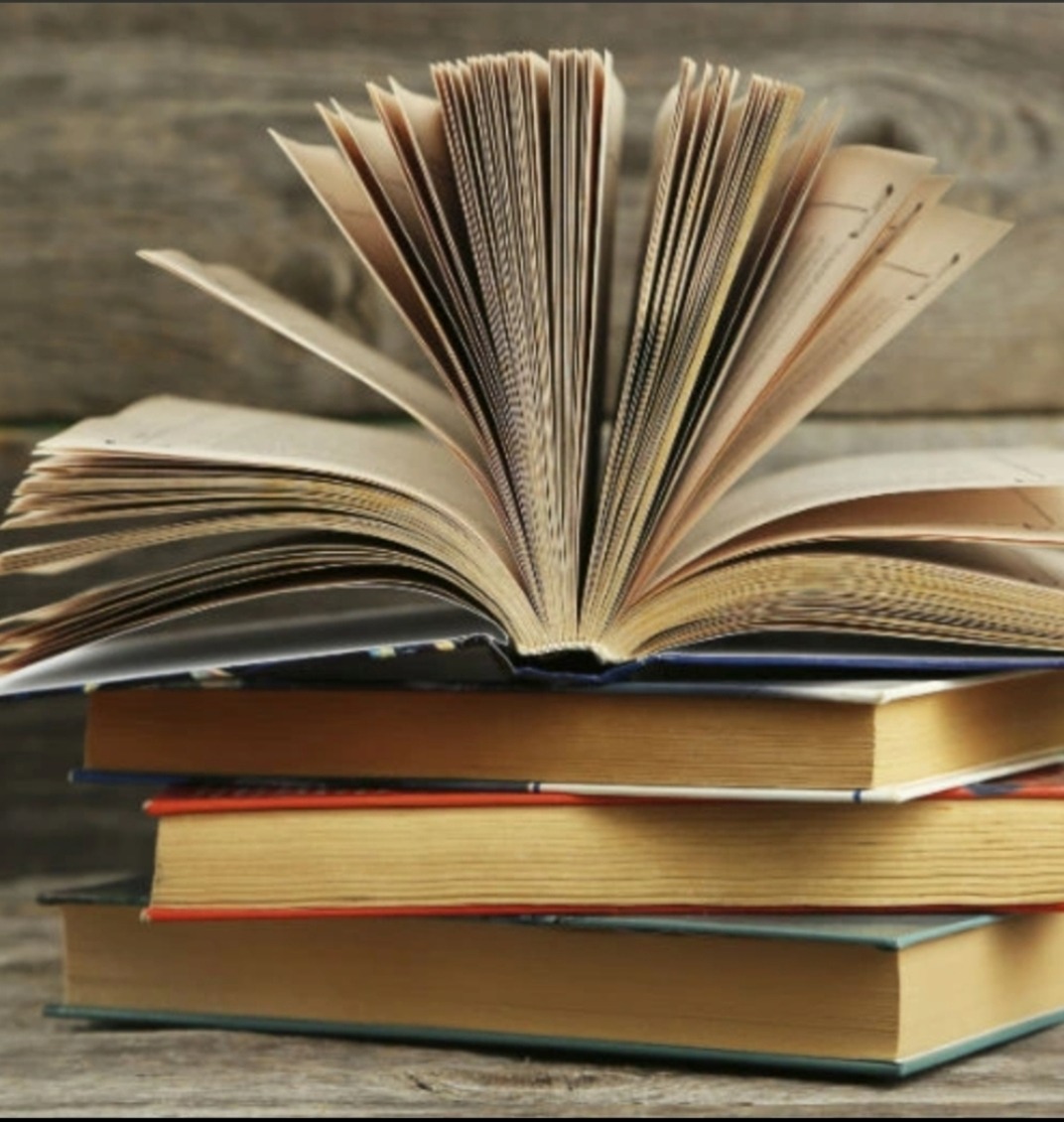 Stos starych książek z otwartą książką na górze, pokazującą strony z tekstem, na tle drewnianej ściany.