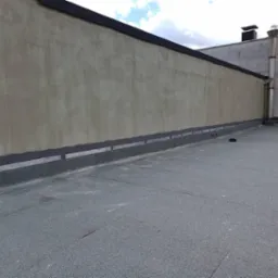 Widok z góry na płaski dach pokryty papą w kolorze szaro-zielonym, z widocznym metalowym obrzeżem przy ścianie i kominem w tle.