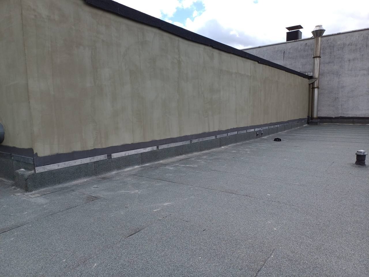 Widok z góry na płaski dach pokryty papą w kolorze szaro-zielonym, z widocznym metalowym obrzeżem przy ścianie i kominem w tle.