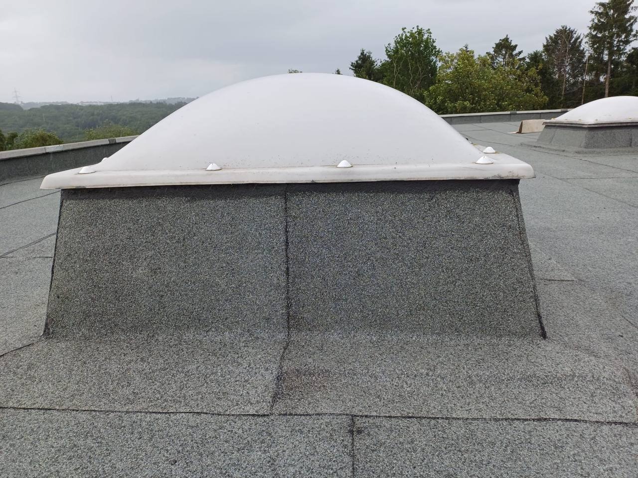 Płaski dach pokryty papą z zamontowanym świetlikiem kopułowym, widok z góry na tle zieleni