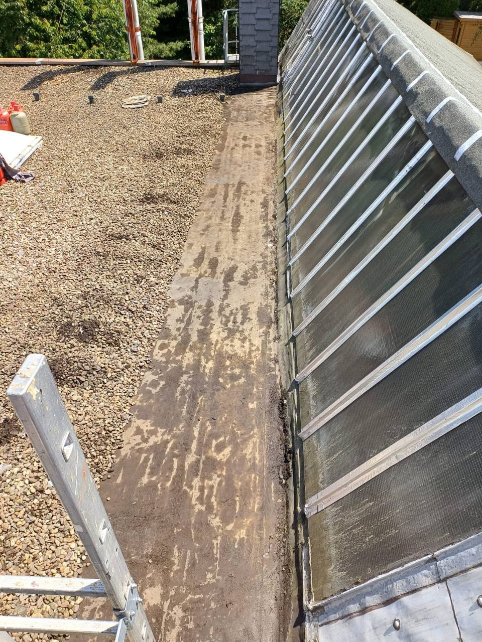 Fragment dachu z papy z widocznymi śladami wcześniejszych napraw, obok konstrukcja szklarni z metalowymi ramami, widok z góry z częściowo widoczną drabiną.