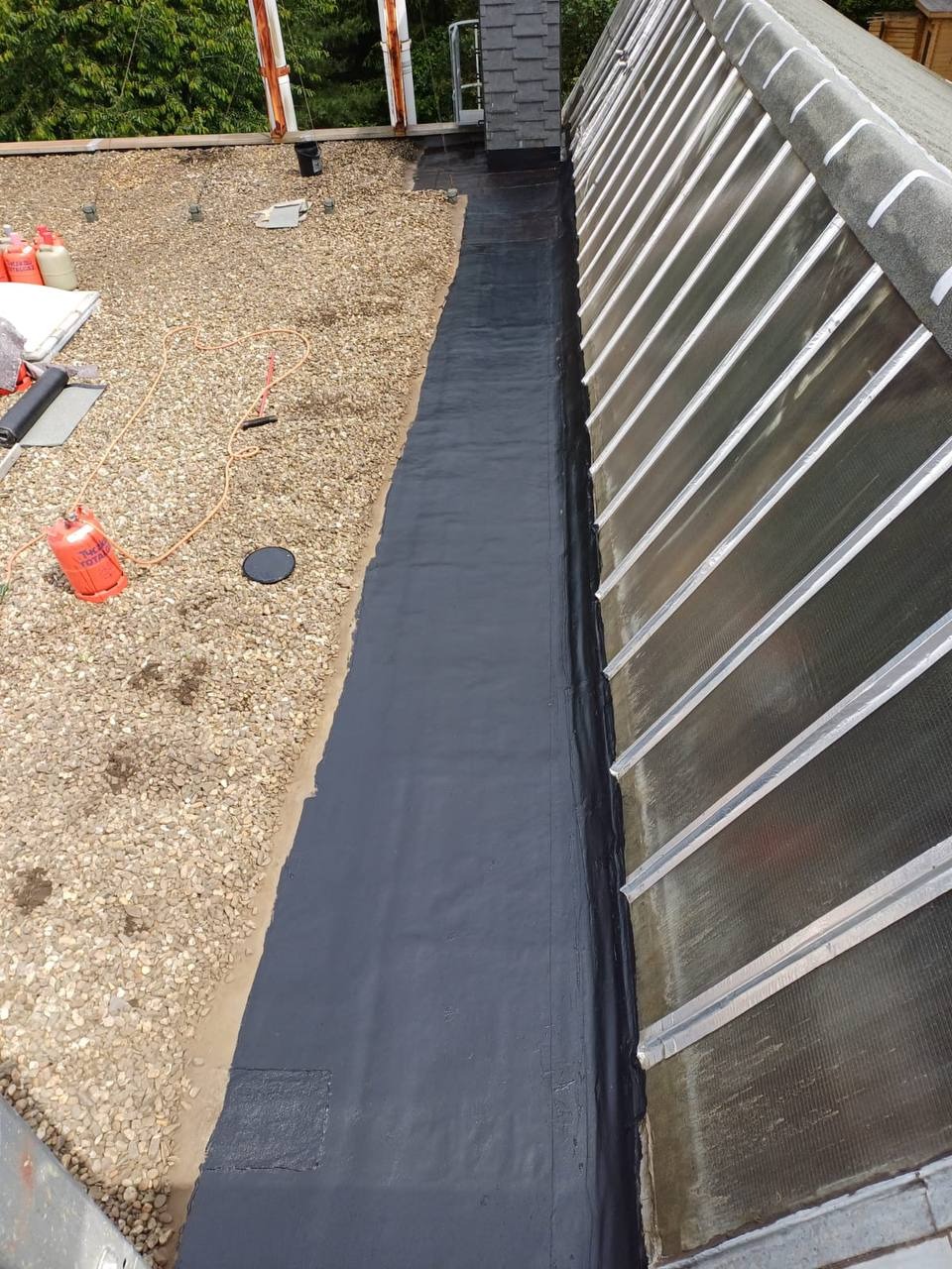 Płaski dach pokryty żwirem z fragmentem uszczelnionej powierzchni bitumicznej, widoczne butle gazowe i narzędzia dekarskie.