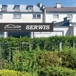 Budynek serwisu samochodowego Ford Jaran z zielonym żywopłotem i metalową bramą w słoneczny dzień. Widoczny szyld reklamowy.