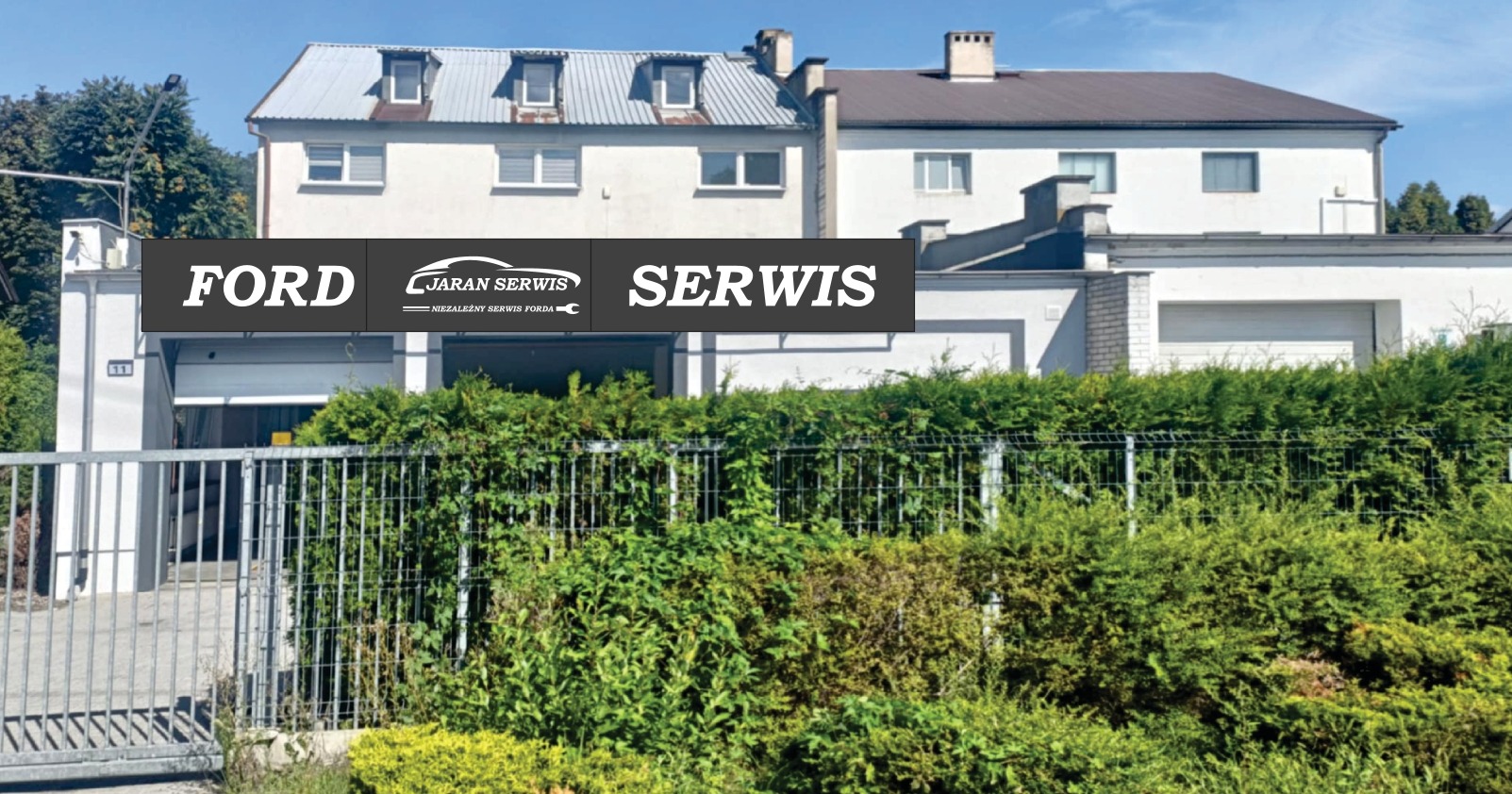 Budynek serwisu samochodowego Ford Jaran z zielonym żywopłotem i metalową bramą w słoneczny dzień. Widoczny szyld reklamowy.