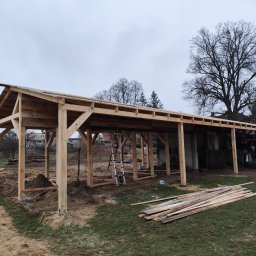 Aurelian Hryszko GOLD BUILD - Wznoszenie drewnianej konstrukcji z widoczną więźbą dachową, drabina oparta o konstrukcję, deski leżące na ziemi, w tle budynki mieszkalne i drzewa bez liści, pochmurne niebo.