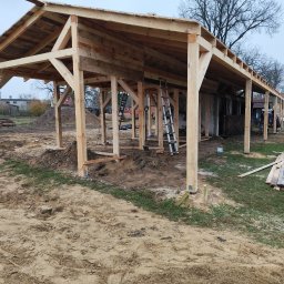 Aurelian Hryszko GOLD BUILD - Konstrukcja drewniana w trakcie budowy, widoczne słupy, więźba dachowa, drabina oparta o konstrukcję, na ziemi leżą deski, w tle budynki mieszkalne i drzewa.