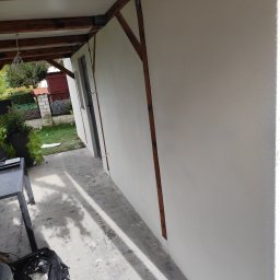 Aurelian Hryszko GOLD BUILD - Świeżo otynkowana ściana zewnętrzna z drewnianą konstrukcją zadaszenia, widoczny fragment terenu zielonego i ślady po pracy budowlanej na posadzce.