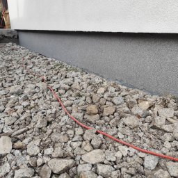 Aurelian Hryszko GOLD BUILD - Przygotowanie terenu pod elewację budynku: widoczne kamienie, kabel i fragment ocieplonej ściany.
