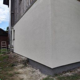 Aurelian Hryszko GOLD BUILD - Fragment ocieplonej elewacji budynku z widoczną fakturą tynku, rurą odprowadzającą spaliny i drabiną opartą o ścianę.