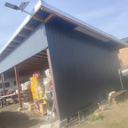 Aurelian Hryszko GOLD BUILD - Zadaszona wiata z czarną blachą trapezową, widoczna konstrukcja drewniana dachu, wewnątrz nagromadzone kartony i przedmioty, na górze lampa solarna.
