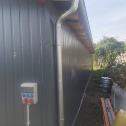 Aurelian Hryszko GOLD BUILD - Szara blaszana ściana budynku gospodarczego z zamontowaną rynną, skrzynką elektryczną z gniazdami siłowymi i widoczną konstrukcją dachu z drewnianych belek.
