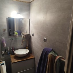 Aurelian Hryszko GOLD BUILD - Szara łazienka z umywalką nablatową na drewnianym blacie, lustro z ornamentem, baterią w kolorze złotym i szarymi płytkami na ścianach.