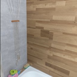 Aurelian Hryszko GOLD BUILD - Szczegółowe ujęcie wykończenia łazienki z drewnopodobnymi płytkami na ścianie, białą wanną, suszarką sznurkową i zabawkami dla dzieci na brzegu wanny.