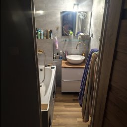 Aurelian Hryszko GOLD BUILD - Wnętrze wykończonej łazienki z wanną, umywalką nablatową na szafce z drewnianym blatem, złotą armaturą i szarą mozaiką na ścianach, widok zza uchylonych drzwi.