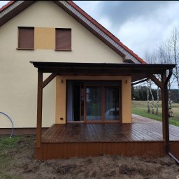 Aurelian Hryszko GOLD BUILD - Nowoczesny taras drewniany z zadaszeniem, przylegający do budynku z brązowymi roletami, widok na brzozy w tle.