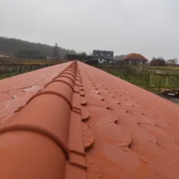 Nowy dach z czerwonej dachówki ceramicznej w kształcie rybiej łuski, pokryty kroplami deszczu, widok z góry na tle zabudowań i zalesionego terenu w pochmurny dzień.