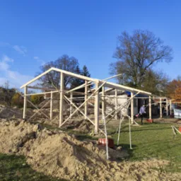 Wznoszona drewniana konstrukcja z widoczną więźbą dachową na tle błękitnego nieba, z hałdami ziemi w pobliżu i pracownikiem w tle.