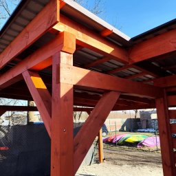 Aurelian Hryszko GOLD BUILD - Konstrukcja drewniana z metalowym zadaszeniem, widok z dołu na połączenie belek i słupów, w tle kolorowe kajaki i ogrodzenie z siatki.