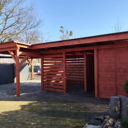Aurelian Hryszko GOLD BUILD - Drewniana konstrukcja zadaszona, częściowo zabudowana panelami z poziomych listew, pomalowana na czerwono-brązowy kolor, z widoczną konstrukcją dachu, na tle trawnika i drzew bez liści.
