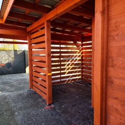 Aurelian Hryszko GOLD BUILD - Nowo wybudowana, pomalowana na brązowo drewniana konstrukcja z metalowym dachem, z poziomymi listwami po bokach, stojąca na szarej kostce brukowej.