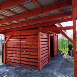 Aurelian Hryszko GOLD BUILD - Drewniana konstrukcja z zadaszeniem z blachy trapezowej, pomalowana na pomarańczowo-czerwony kolor, z widocznymi poziomymi deskami tworzącymi ściany boczne, na tle zielonego żywopłotu i błękitnego...