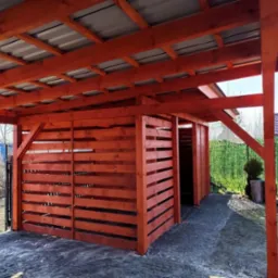 Drewniana konstrukcja z zadaszeniem z blachy trapezowej, pomalowana na pomarańczowo-czerwony kolor, z widocznymi poziomymi deskami tworzącymi ściany boczne, na tle zielonego żywopłotu i błękitnego...