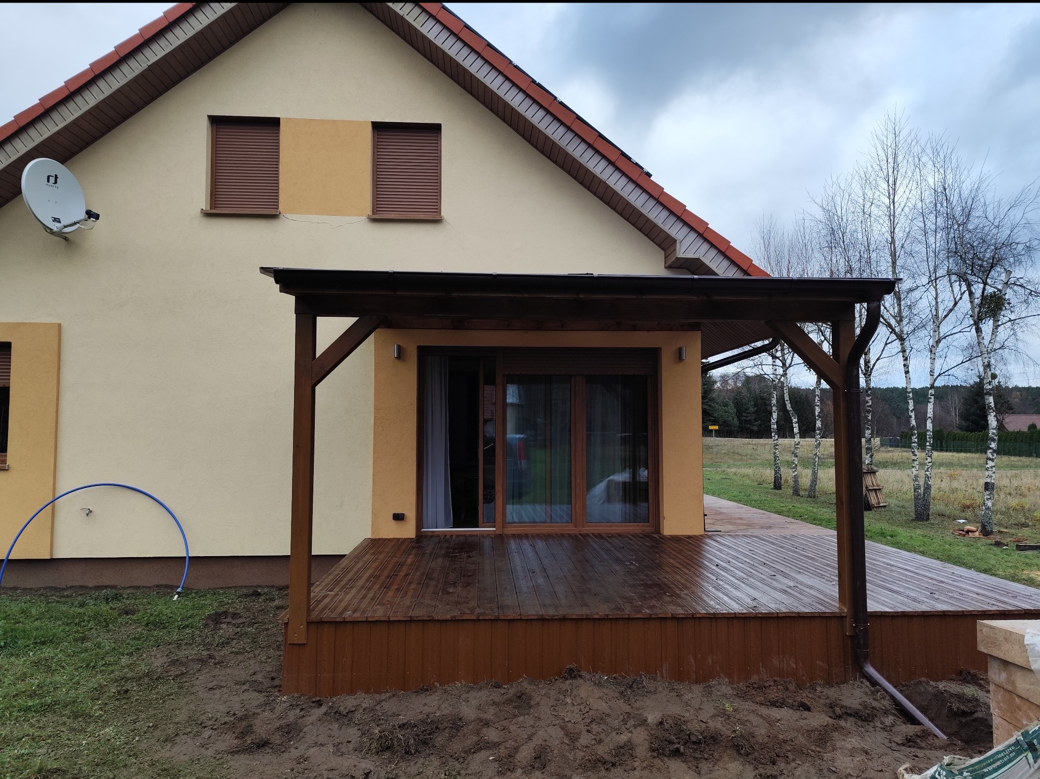 Nowoczesny taras drewniany z zadaszeniem, przylegający do budynku z brązowymi roletami, widok na brzozy w tle.