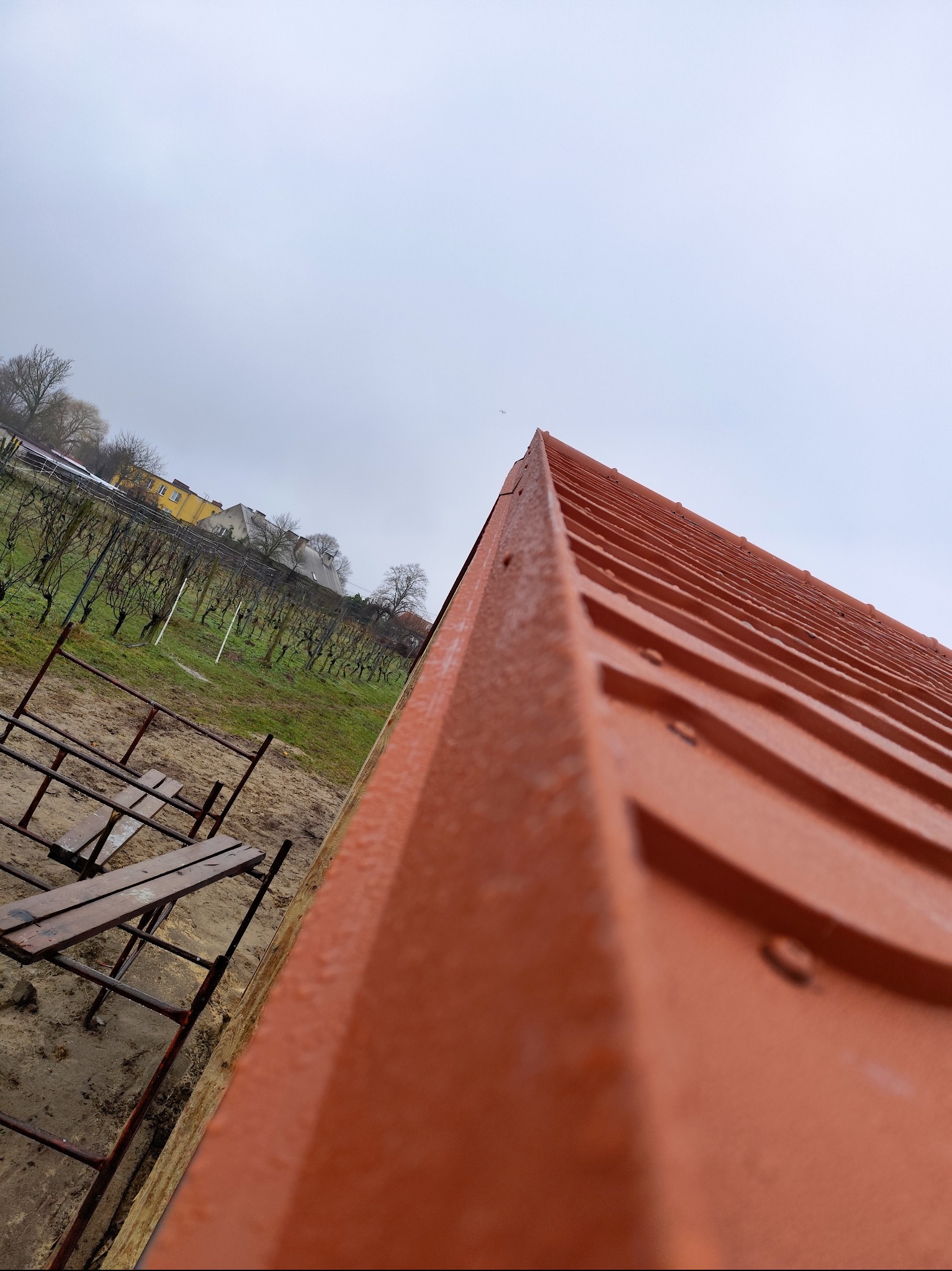 Fragment pomarańczowego dachu z metalowej dachówki widziany z dołu, w tle rusztowanie, winnica i zabudowania w pochmurny dzień.