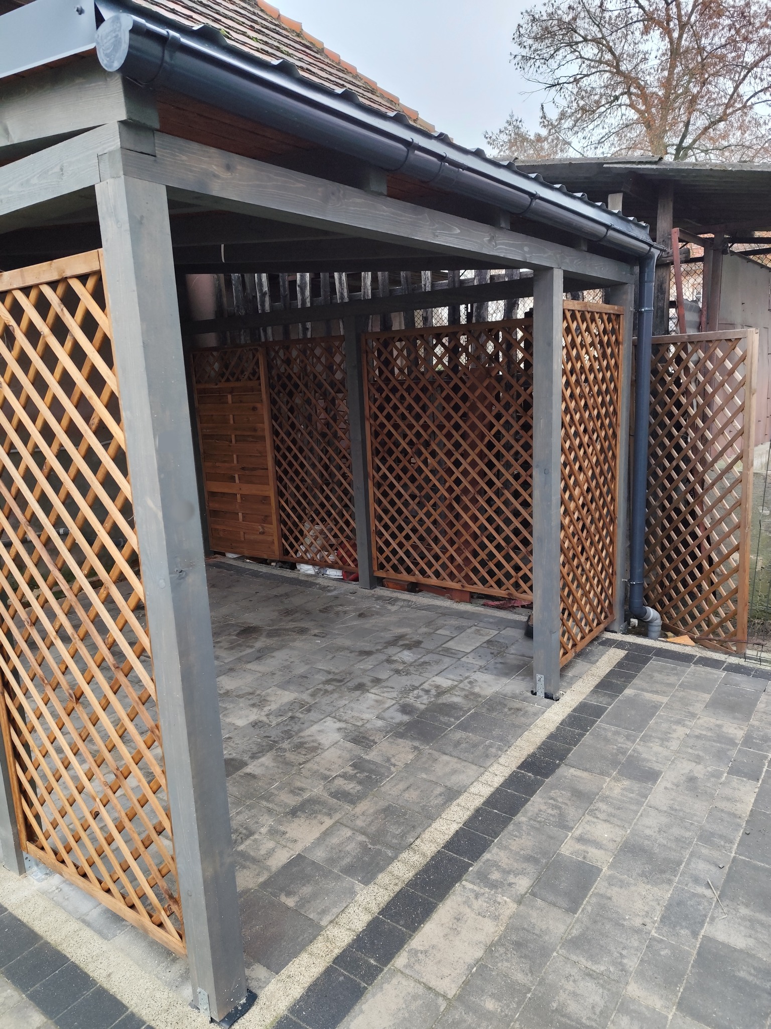 Szara drewniana konstrukcja altany z kratownicowymi ścianami, widok z zewnątrz na kostkę brukową i fragment dachu domu w tle.