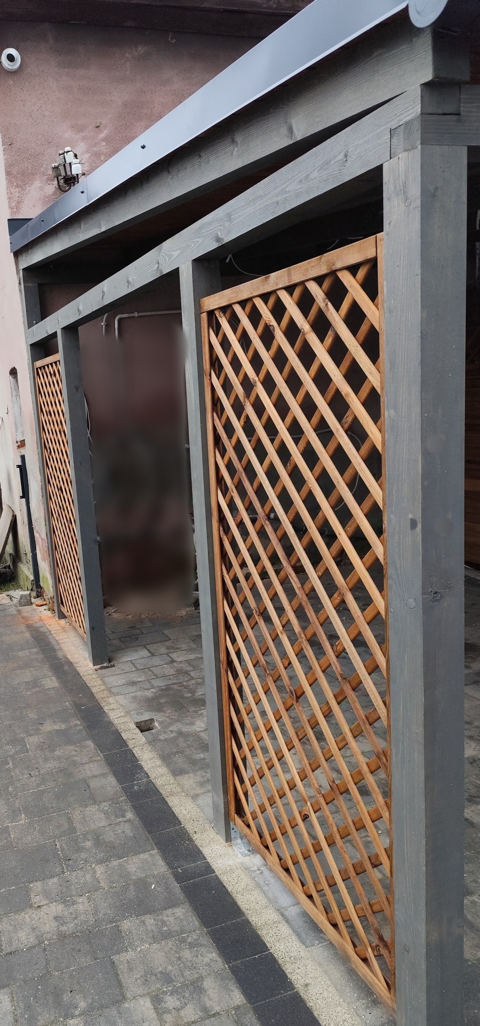 Konstrukcja z drewnianymi kratami, prawdopodobnie fragment zadaszenia tarasu lub altany, z szarą ramą, widoczna z poziomu chodnika wyłożonego kostką.