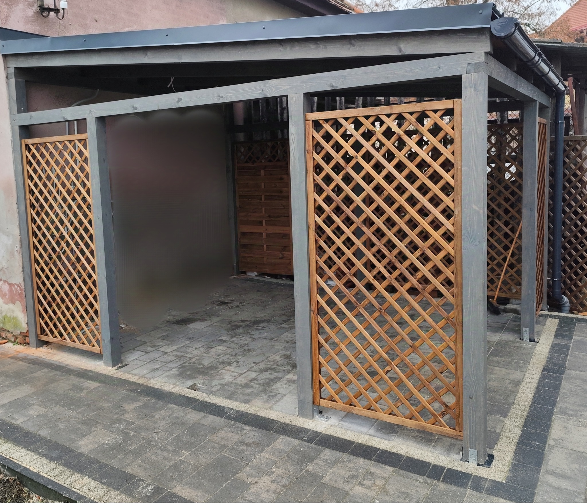 Zadaszona konstrukcja drewniana z ażurowymi ściankami, pomalowana na szaro, z widocznym fragmentem brukowanego podłoża.