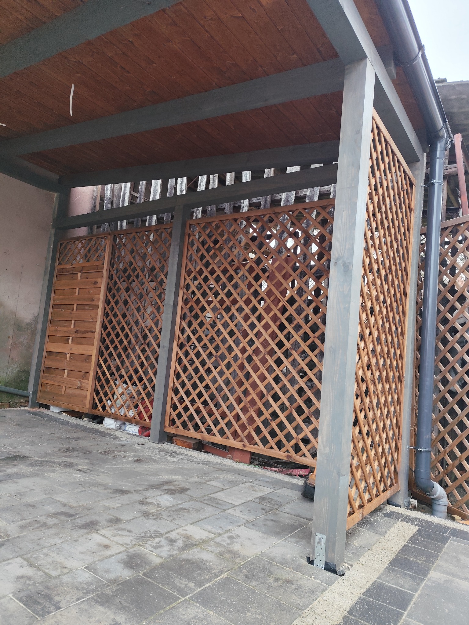 Drewniana altana z kratownicą i zadaszeniem z desek, szare słupy nośne, widoczne rynny i kostka brukowa.