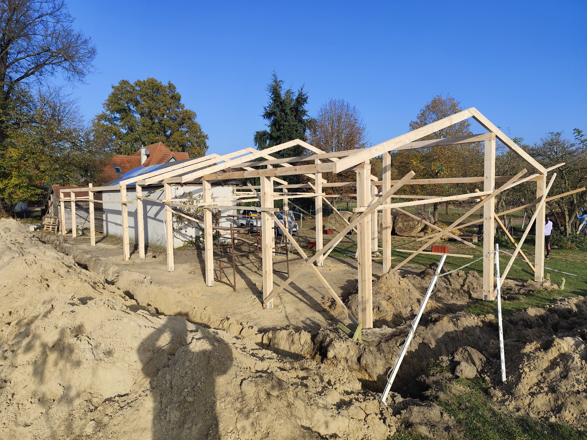 Konstrukcja drewniana w trakcie budowy, widoczne słupy i elementy szkieletu dachu na tle błękitnego nieba, ziemia wykopana wokół fundamentów.