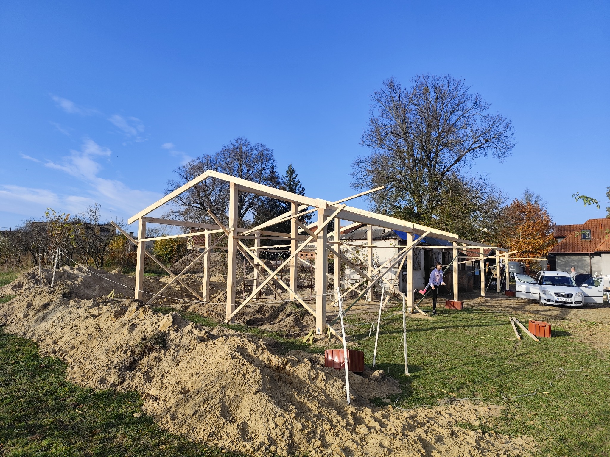 Wznoszona drewniana konstrukcja z widoczną więźbą dachową na tle błękitnego nieba, z hałdami ziemi w pobliżu i pracownikiem w tle.