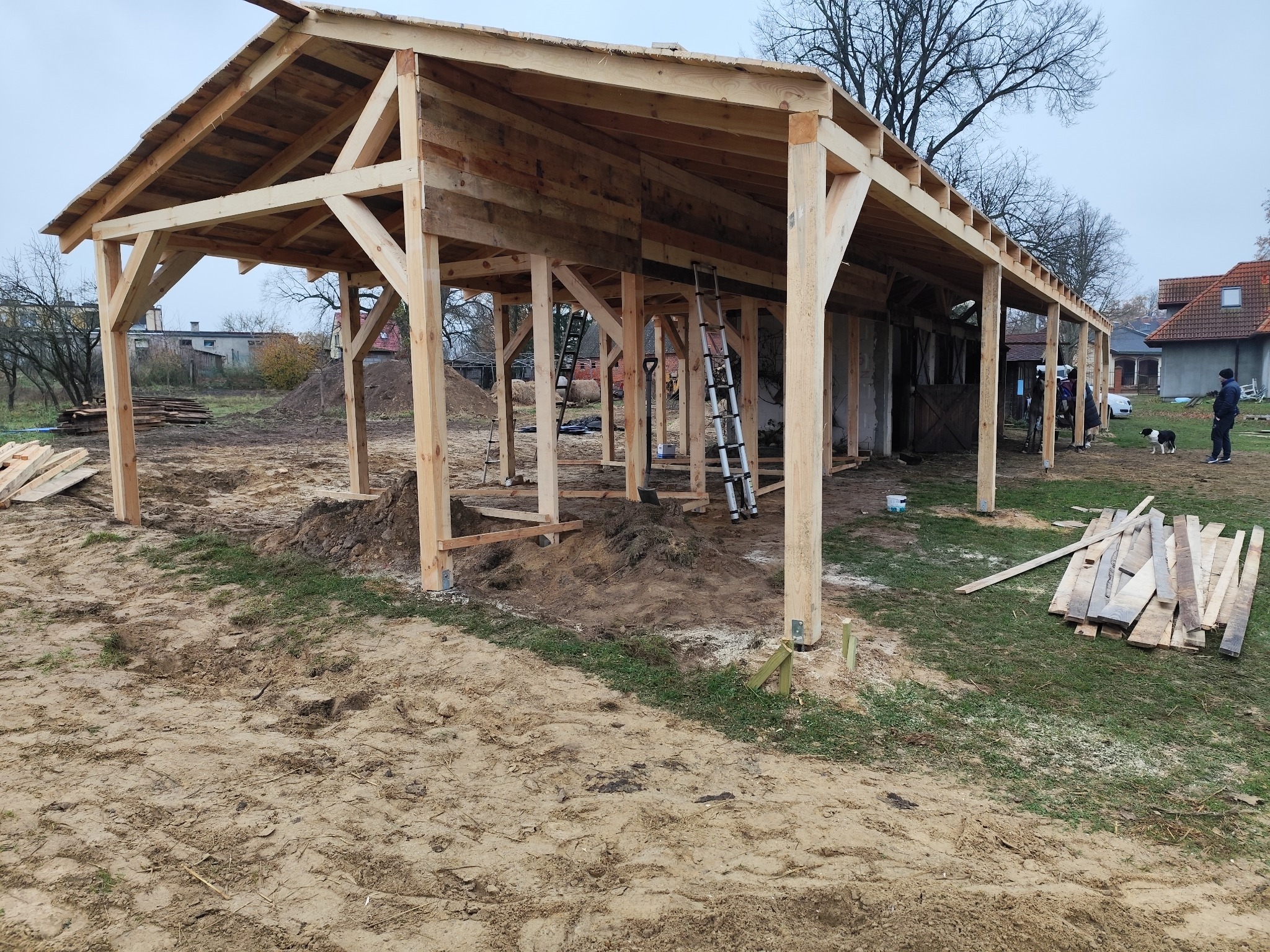 Konstrukcja drewnianej altany w trakcie budowy, widoczne słupy, więźba dachowa i elementy dachu, na ziemi leżą deski, w tle domy i drzewa.