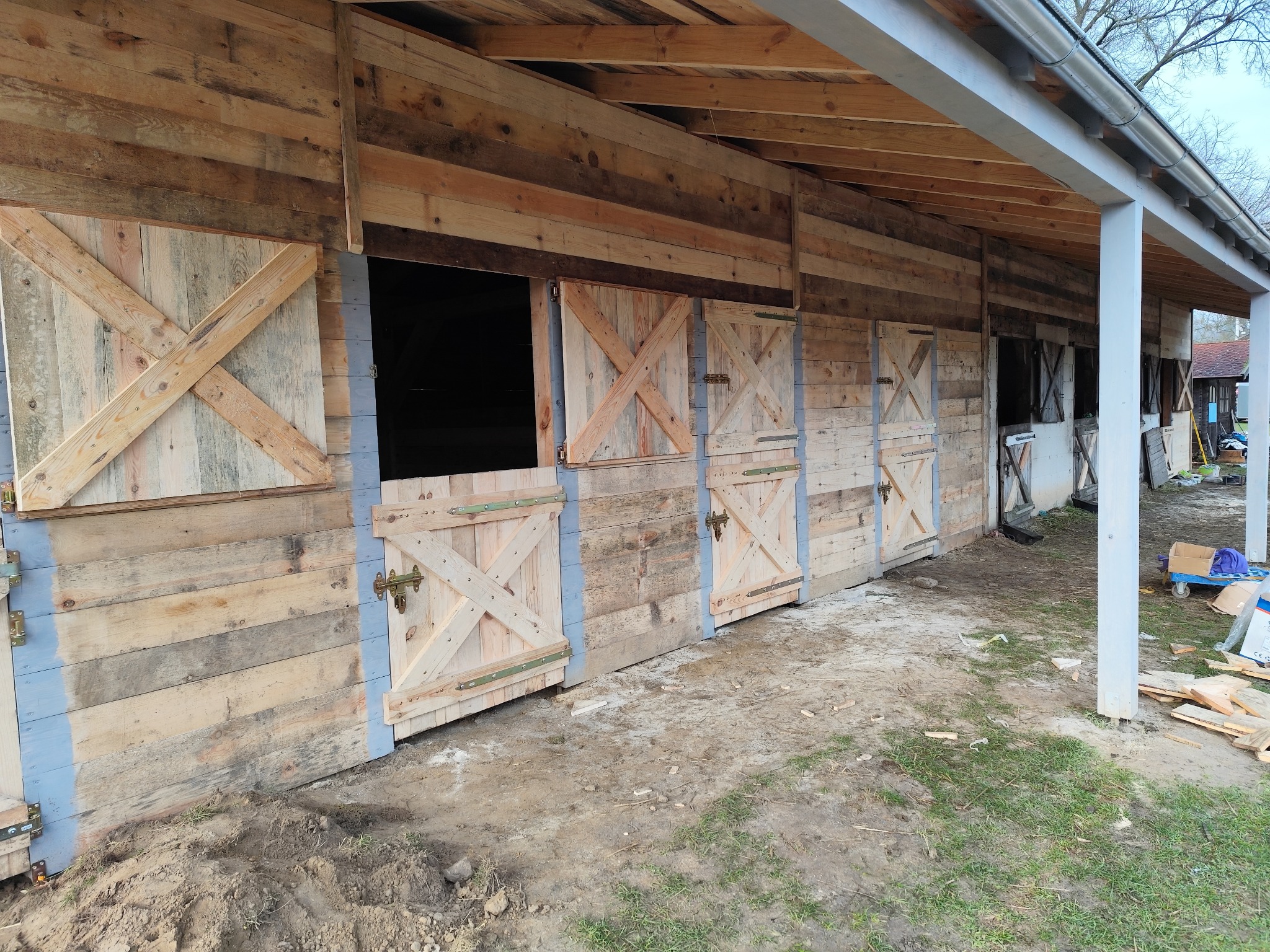 Drewniana konstrukcja z otwieranymi okiennicami w kształcie litery X, wykonana z surowego drewna, z widocznymi śladami obróbki i szarą farbą na krawędziach, pod zadaszeniem z drewnianą więźbą dachową.