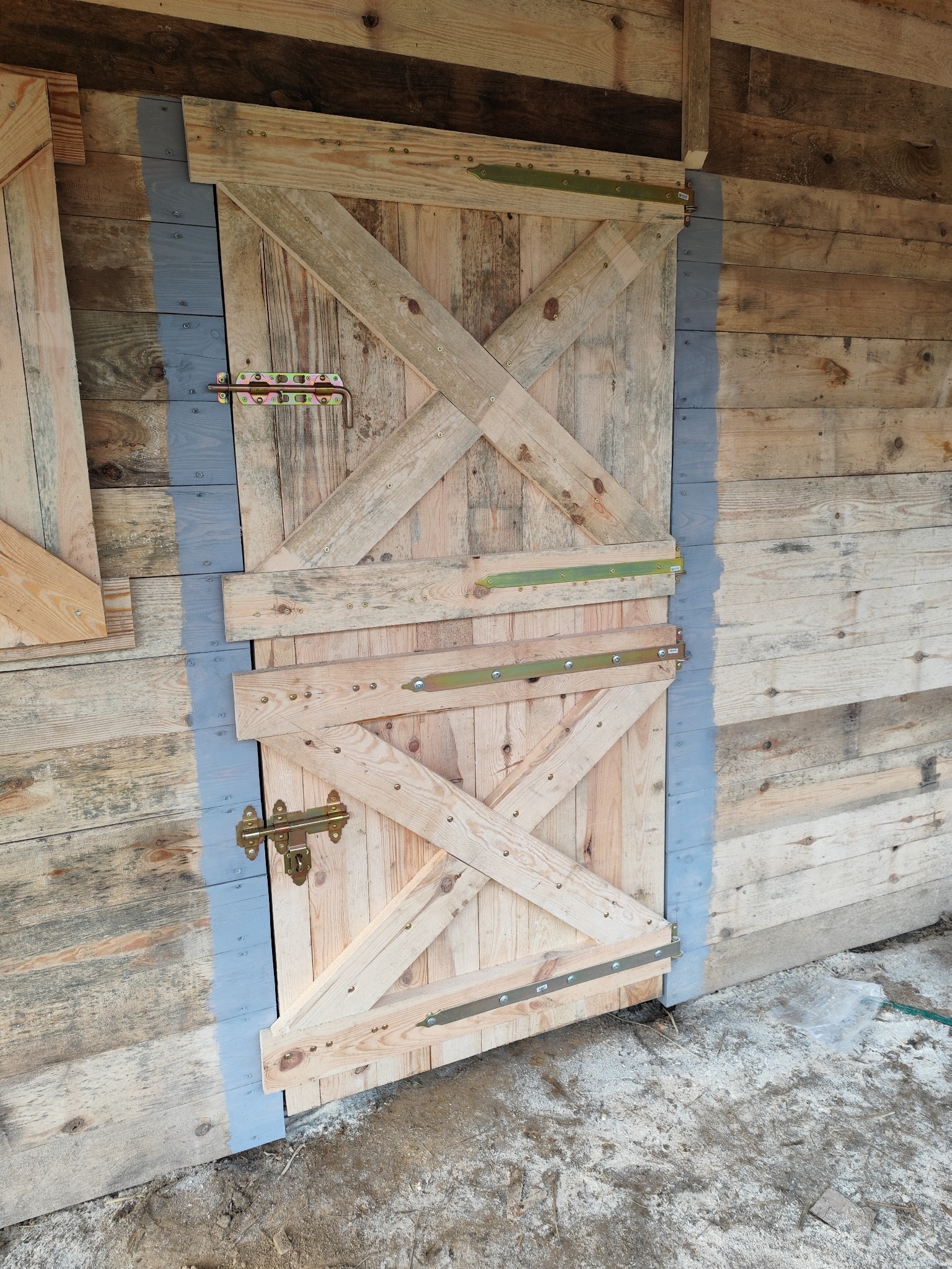 Drewniana brama z ozdobnym krzyżowym wzmocnieniem, zamontowana na drewnianej konstrukcji, z widocznymi zawiasami i zasuwą, obok ściana z desek pomalowana na szaro.