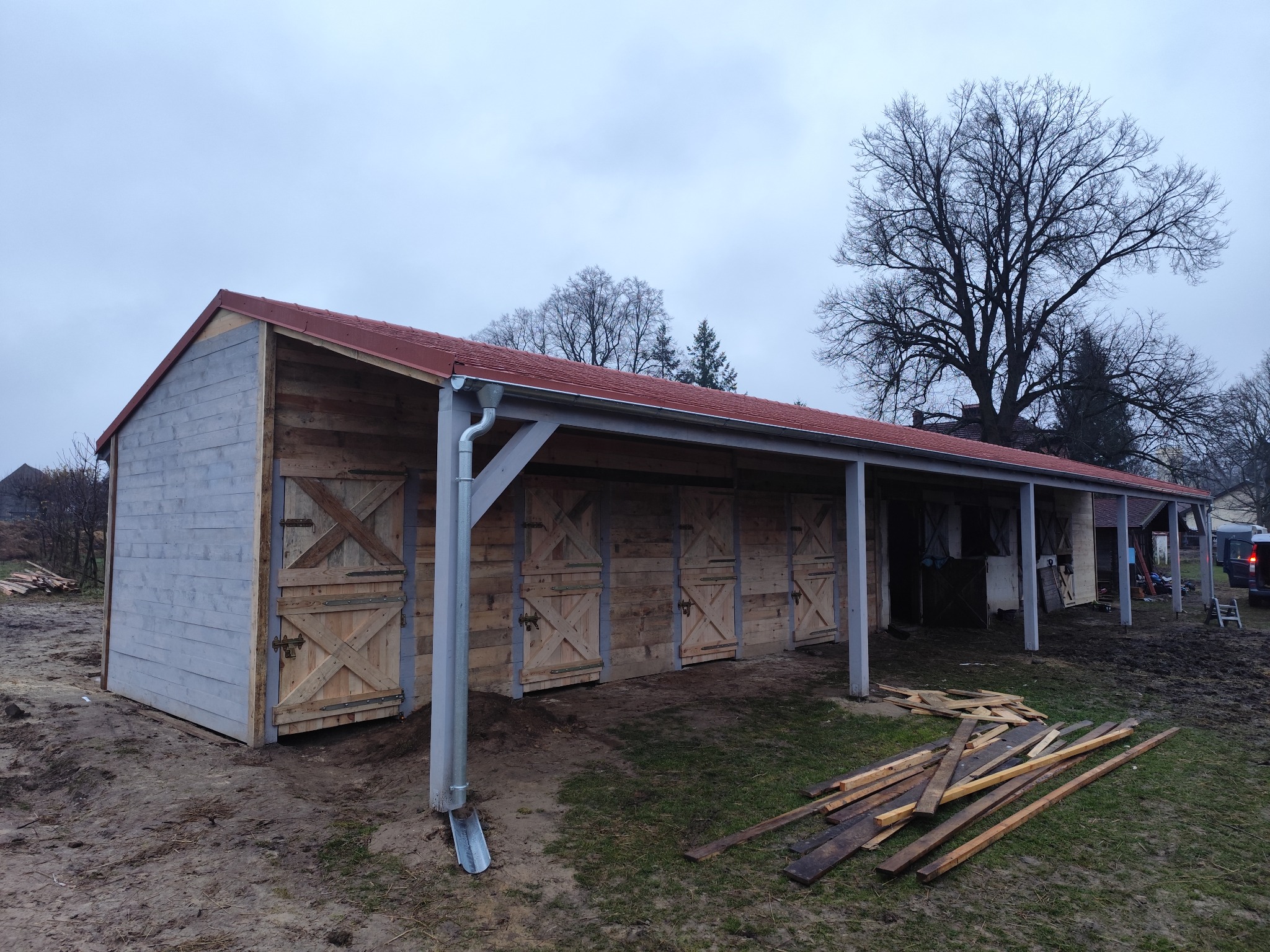 Budowa drewnianej konstrukcji z czerwonym dachem i szarymi elementami, widoczne drewniane drzwi z krzyżowym wzorem, rynna, oraz stos desek na ziemi.